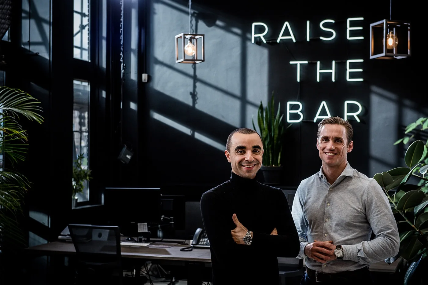 twee mannen voor een muur waar groot Raise the bar staat