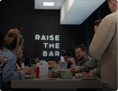 Collega's eten Lunch met grote tekst Raise the Bar op de muur